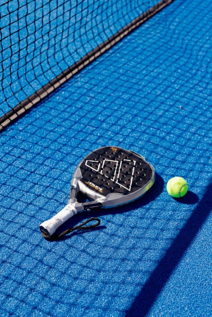 Padel racket and ball resting on a vibrant blue court, next to a net, under bright daylight.