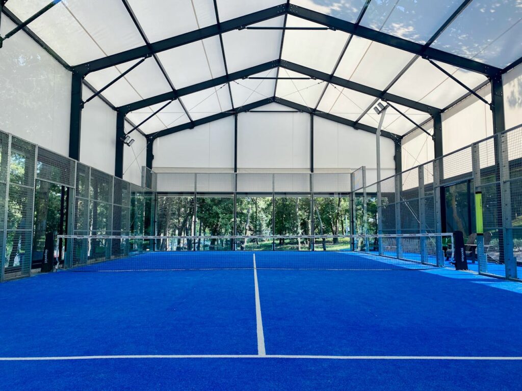 Spacious indoor padel court with vibrant blue flooring under a wide roof.